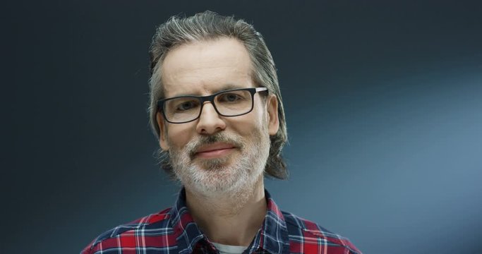 Close up of Caucasian midde-aged handsome man in motley shirt and with gray beard touching glasses and turning face while looking to camera with smile on grey wall background. Portrait of male.