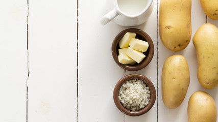 Raw potatoes on a right side on a white wooden background. There is salt, butter in brown bowls and milk near it. 