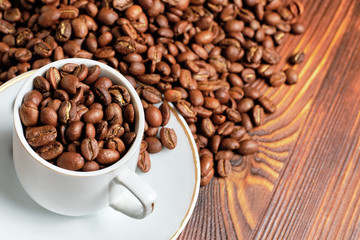 Coffee roasted grains are scattered in a white cup and on a wooden brown background.