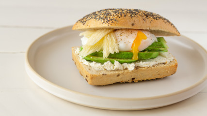 Avocado, soft cheese, poached egg sandwich lying on a round plate on a right side. Egg yolk flow out. Wooden white background. 