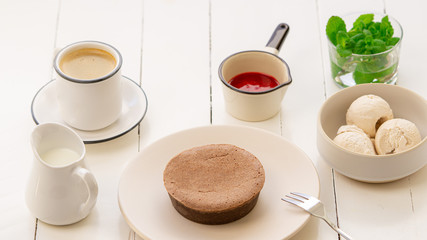 Chocolate cake on a round plate. There is a bowl with ice cream, mint and strawberry sauce near it. Also there is a coffee cup and jar of milk
