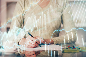 Hand taking notes in notepad. Forex chart holograms in front. Concept of research. Double exposure