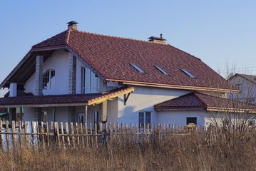 Obraz premium large white private house under a brown tiled roof with windows against a blue sky behind a wooden fence in dry gray grass