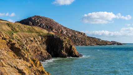 Fototapeta premium Irish landscape with rugged cliffs and blue sea. Part of the Howth Cliff Walk path with the Red Rock peak in the distance, near Sutton, Dublin, Ireland.