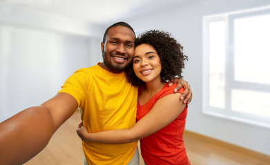 Fototapeta na wymiar new home, real estate and people concept - happy smiling african american couple taking selfie over empty apartment on background