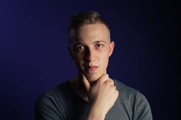 Young caucasian blonde man in grey t shirt on dark purple background touching his chin and thinking about something