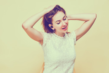 A girl in summer clothes stands on a yellow background and listens to music in wireless headphones and drinks while tucking her hair into a ponytail. Toned.