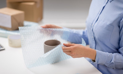 delivery, mail service, people and shipment concept - close up of woman packing mug and it wrapping into protective bubble wrap at post office