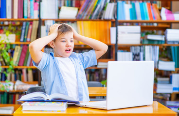 A boy sitting at a table in the library at a laptop, looking at the screen in surprise and folded his hands on his head