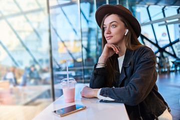 Portrait of stylish trendy attractive hipster casual female in hat with wireless headphones in cafe. Modern people with technology digital lifestyle