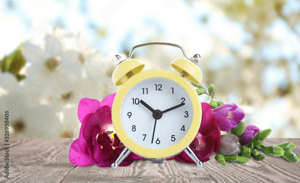Poster Yellow alarm clock and flowers on table against blurred background. Spring time