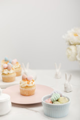Obraz na płótnie Canvas Selective focus of cupcakes with decorative bunnies, bowl with meringues and flowers on grey background