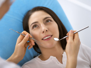 Smiling brunette woman being examined by dentist at dental clinic. Hands of a doctor holding dental instruments near patient's mouth. Healthy teeth and medicine concept