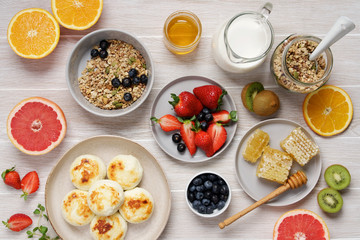 Healthy breakfast over white wooden table. Variety of fruits, granola, honey, milk and curd pancakes