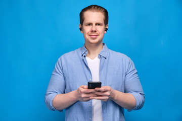 A man in casual clothes on a blue background holds a smartphone in his hands and looks into the camera in his wireless headphones in his ears.