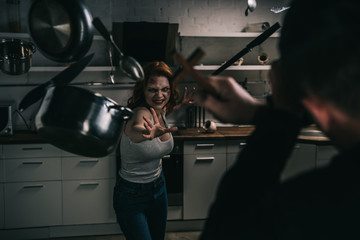 selective focus of creepy demon with levitating cookware and exorcist with cross in kitchen