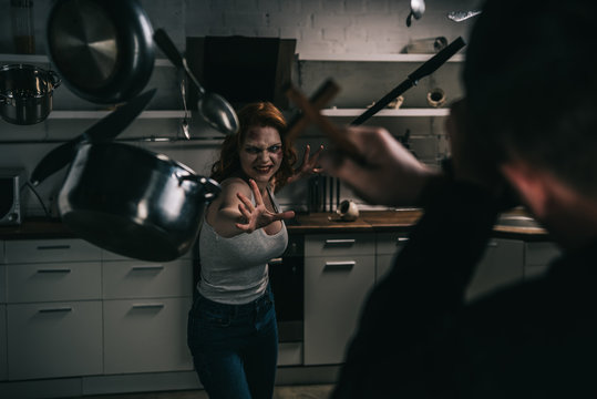 selective focus of creepy demon with levitating cookware and exorcist with cross in kitchen