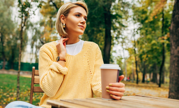 Image of attractive young woman drinking coffee at the terrace cafetaria in the park. Blonde female in the city street drinking a hot beverage, looking at one side. Pretty girl with a cup of coffee.