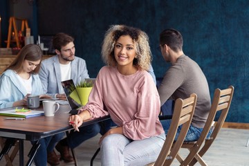 Happy to be in team. Beautiful cheerful African woman looking at camera with smile while sitting at the office table with her coworkers.