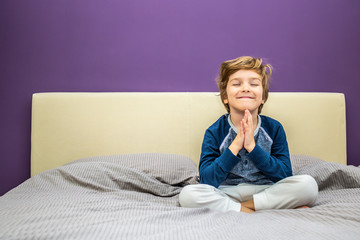 Happy boy with hands clasped meditating on the bed.