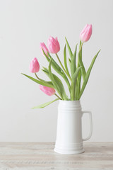 pink tulips in white ceramic jug on wooden table on background white wall