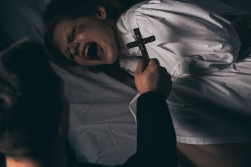 exorcist holding cross over demonic yelling girl in bed - obrazy, fototapety, plakaty
