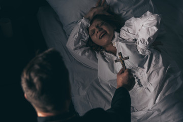 exorcist holding cross over demonic screaming girl in bed