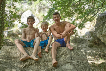 Three brothers having fun making faces and enjoying their time in summer happily smiling and relaxed sitting in shorts on a rock