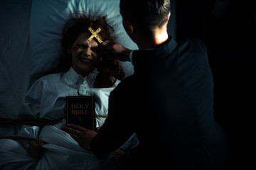 exorcist with bible and cross standing over demonic obsessed girl in bed - obrazy, fototapety, plakaty