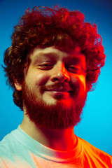 Obraz na płótnie Canvas Calm, delighted. Caucasian close up man's portrait isolated on blue studio background in red neon light. Beautiful male model, red curly hair. Concept of human emotions, facial expression, sales, ad.