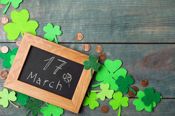 Chalkboard with written 17 March, clover leaves and gold coins on blue wooden background. St....