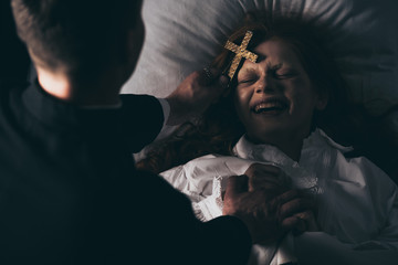 exorcist holding cross over laughing demon in bed - obrazy, fototapety, plakaty