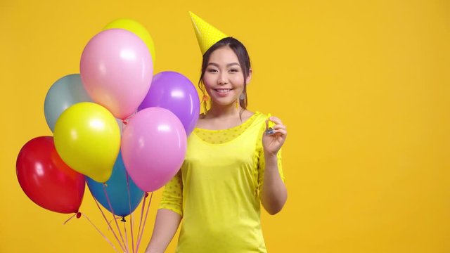 asian girl with balloons blowing party blower isolated on yellow