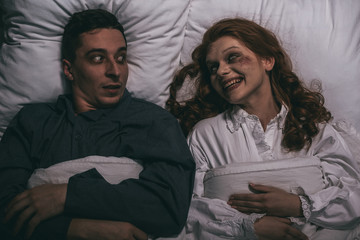 top view of female smiling demon lying in bed with scared man