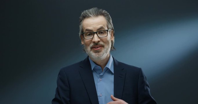 Portrait of rich handsome businessman of middle-age in glasses and suit speaking to camera on grey background. Happy man financial couch in business style taking out and showing money dollars.