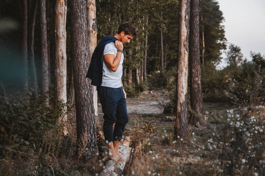Handsome man posing outdoors in forest stands barefoot on a log, wearing checked jacket. Place for text or advertising