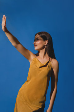 Portrait Of Young Girl Covering Her Face To Block Sun Light. Shadow And Light Over Woman's Face. Creative Portraits. Female In Orange Summer Beach Dress On A Blue Background