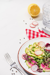 Fototapeta na wymiar selective focus of fresh radish salad with greens and avocado on plate on white surface with water, lemon and cutlery