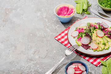 fresh radish salad with greens and avocado served on napkin with cutlery near ingredients in bowls on grey concrete surface