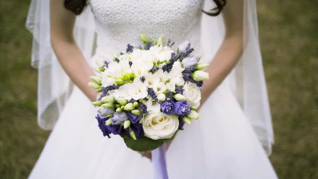 Close up of vivid wedding bouquet at young bride on green grass background, family and celebration concept. Action. Slim woman in white dress holding beautiful wedding bouquet of purple and white