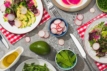 top view of fresh radish salad with greens and avocado served on plates on grey concrete surface with ingredients, cutlery and napkins