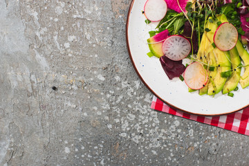 top view of fresh radish salad with greens and avocado on grey concrete surface with fork and napkin