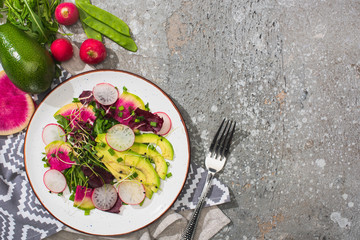 top view of fresh radish salad with greens and avocado near fork and vegetables on grey concrete surface