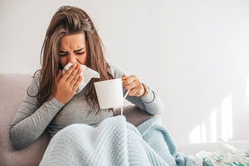 Sick woman blowing her nose. Sick young woman at home on the sofa with a cold, she is covering with a blanket and blowing her nose. Woman drinking hot tea and feeling comfortable