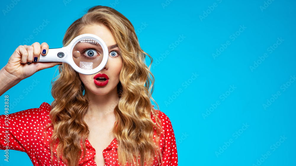 Wall mural surprised blonde holds a magnifying glass in front eye. wonder young woman holds a magnifying glass 