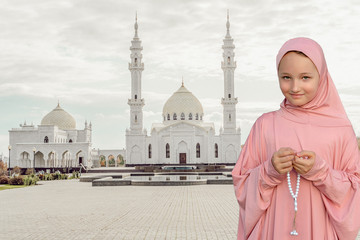 A child in a pink hijab with a beads in his hands Against the background of a white mosque. People...
