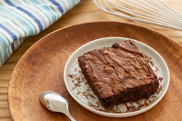 Brownies cake in a light gray plate with grated chocolate and silver spoon in a wooden plate by a white whisk over an oak table background.