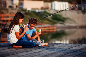 Two happy little friends, boy and girl talking, eating sandwiches and fishing on a lake