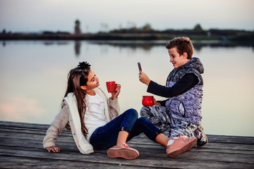 Two young cute little friends, boy and girl  having fun and drinking tea while sitting by the lake in the evening.