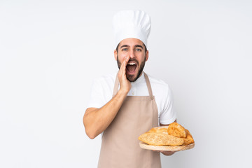 Male baker holding a table with several breads isolated on white background shouting with mouth wide open